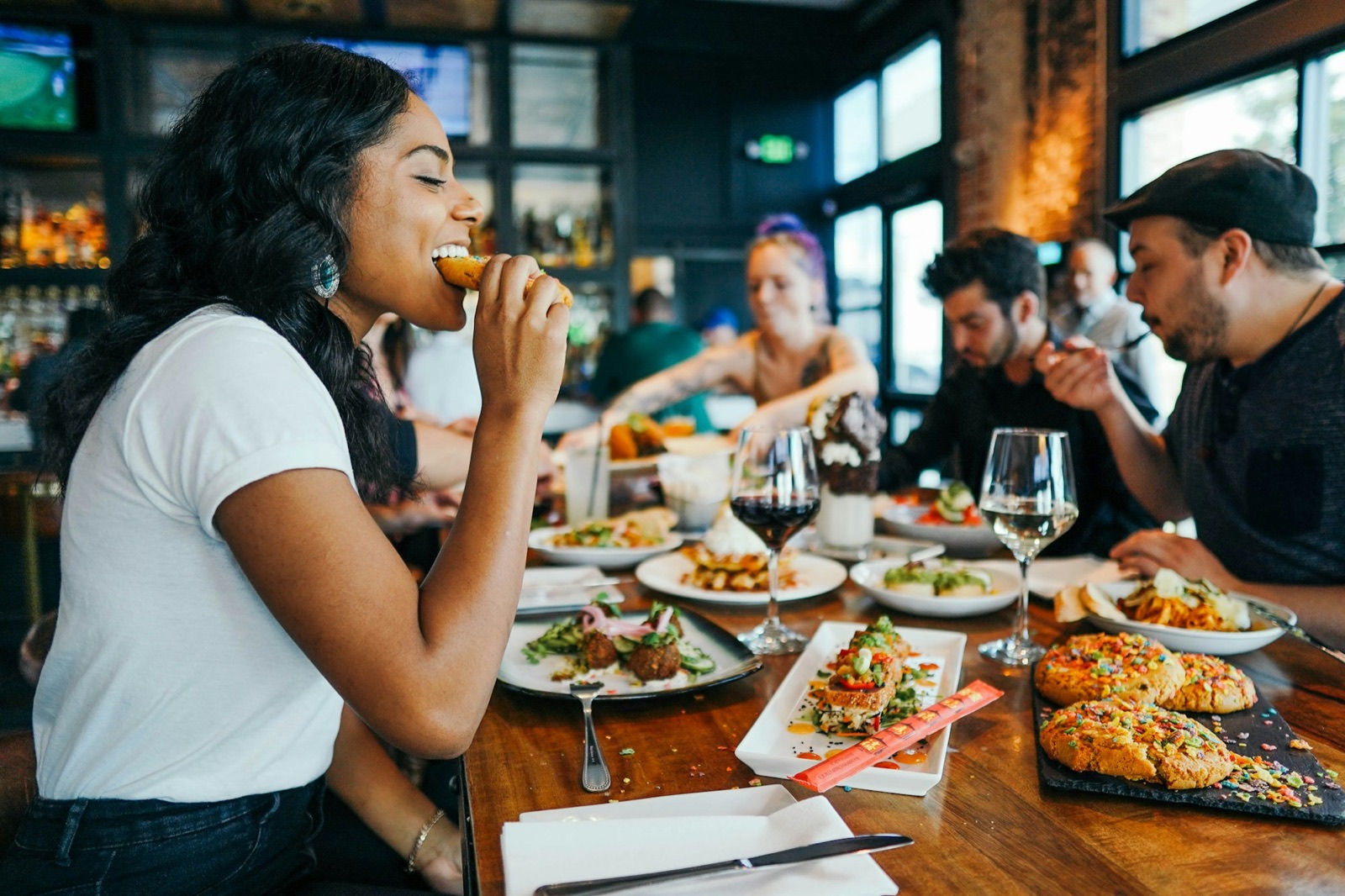 Friends sharing a long dinner table at a local restaurant, one of them laughing mid-bite